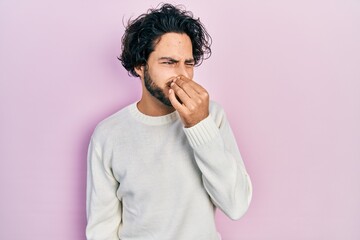 Handsome hispanic man wearing casual white sweater smelling something stinky and disgusting, intolerable smell, holding breath with fingers on nose. bad smell