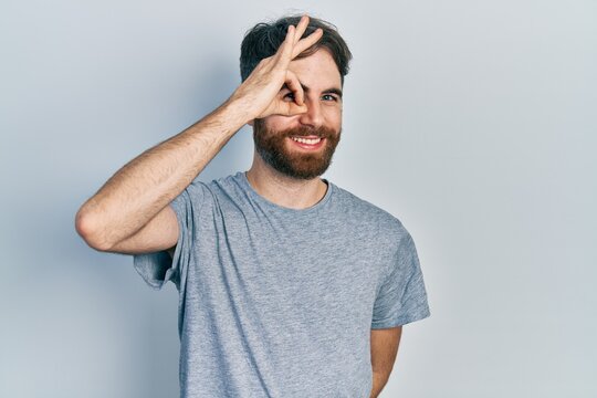 Caucasian man with beard wearing casual grey t shirt doing ok gesture with hand smiling, eye looking through fingers with happy face.