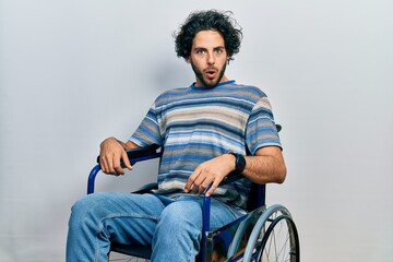 Handsome hispanic man sitting on wheelchair in shock face, looking skeptical and sarcastic,...