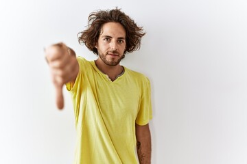 Young hispanic man standing over isolated background looking unhappy and angry showing rejection and negative with thumbs down gesture. bad expression.