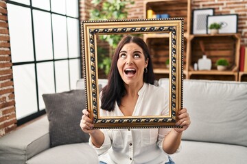 Young hispanic woman holding empty frame angry and mad screaming frustrated and furious, shouting with anger looking up.