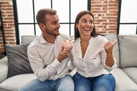 Young hispanic couple holding piggy bank sitting on the sofa pointing thumb up to the side smiling happy with open mouth