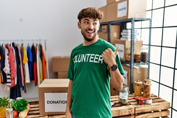 Young arab man wearing volunteer t shirt at donations stand smiling with happy face looking and pointing to the side with thumb up.