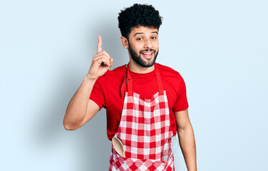 Fototapeta premium Young arab man with beard wearing cook apron pointing finger up with successful idea. exited and happy. number one.