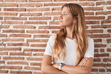 Young caucasian woman standing over bricks wall looking to the side with arms crossed convinced and confident