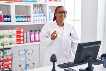 Naklejka na ściany i meble African american woman pharmacist holding pills bottle using computer at pharmacy