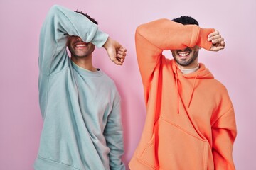 Young hispanic gay couple standing over pink background covering eyes with arm smiling cheerful and...