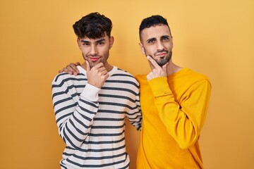 Young hispanic gay couple standing over yellow background looking confident at the camera smiling with crossed arms and hand raised on chin. thinking positive.