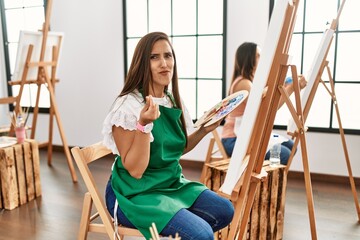 Young hispanic artist women painting on canvas at art studio doing money gesture with hands, asking...