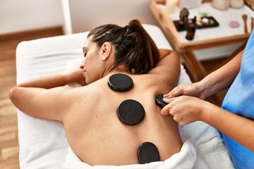 Two women therapist and patient having back massage session using black stones at beauty center