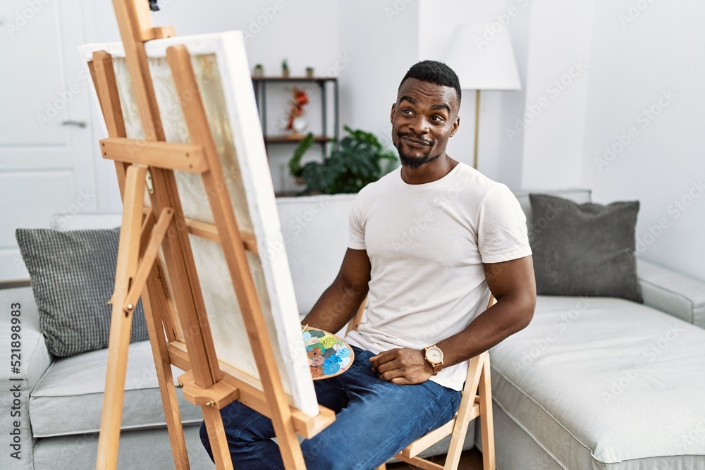 Wall mural Young african man painting on canvas at home smiling looking to the side and staring away thinking.