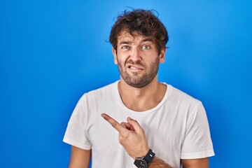 Hispanic young man standing over blue background pointing aside worried and nervous with forefinger, concerned and surprised expression