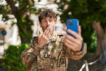Hispanic young man wearing camouflage army uniform doing video call covering mouth with hand, shocked and afraid for mistake. surprised expression