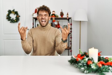Young handsome man with beard sitting on the table by christmas decoration shouting with crazy...