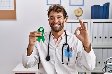 Young hispanic doctor man holding support green ribbon at clinic doing ok sign with fingers, smiling friendly gesturing excellent symbol