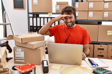 Young hispanic call center agent man working at warehouse very happy and smiling looking far away with hand over head. searching concept.