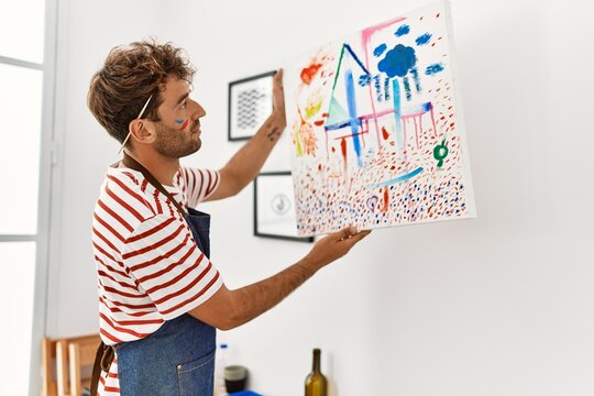 Young hispanic man hanging canvas on wall at art studio