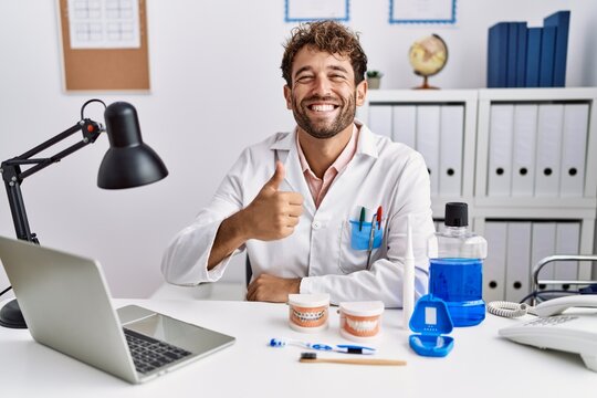 Young hispanic dentist man working at medical clinic doing happy thumbs up gesture with hand. approving expression looking at the camera showing success.