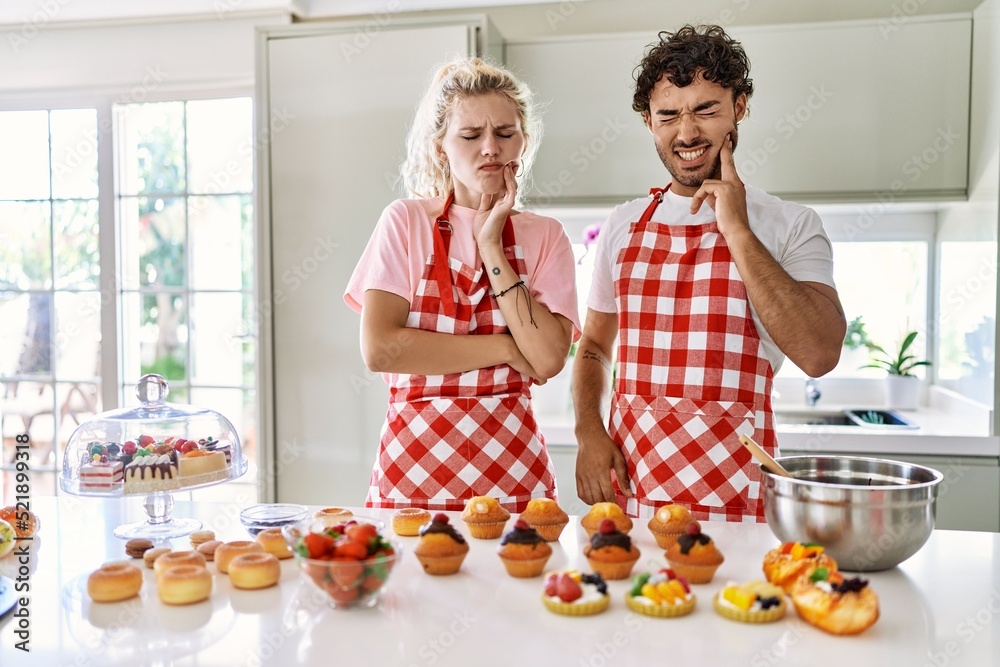 Poster couple of wife and husband cooking pastries at the kitchen touching mouth with hand with painful exp