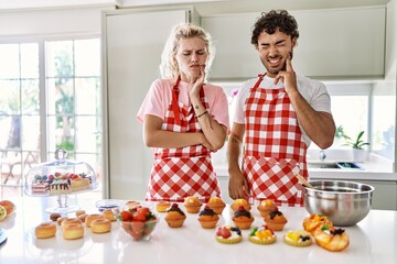 Couple of wife and husband cooking pastries at the kitchen touching mouth with hand with painful expression because of toothache or dental illness on teeth. dentist