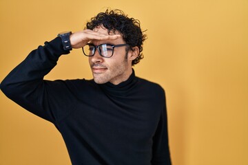 Hispanic man standing over yellow background very happy and smiling looking far away with hand over head. searching concept.