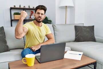 Young man with beard using laptop at home angry and mad raising fist frustrated and furious while shouting with anger. rage and aggressive concept.