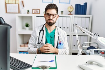 Young man with beard wearing doctor uniform and stethoscope at the clinic puffing cheeks with funny face. mouth inflated with air, crazy expression.