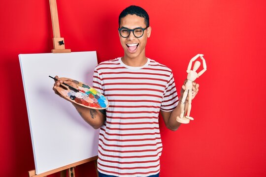 Young african american guy holding painter palette and art manikin winking looking at the camera with sexy expression, cheerful and happy face.