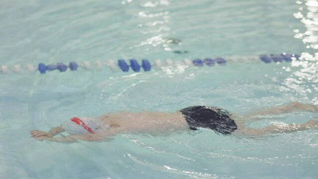 Baby Swimmer Learning To Swim Using Only Legs In Blue Transparent Swimming Pool Water Indoors. Child Kid Boy In Red Swimming Goggles And White Cap Diving, Practicing Swim Exercise Training Workout