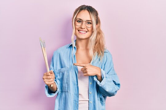 Beautiful young blonde woman holding paintbrushes smiling happy pointing with hand and finger