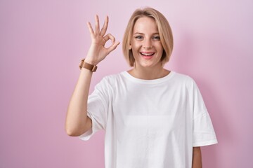 Young caucasian woman standing over pink background smiling positive doing ok sign with hand and fingers. successful expression.