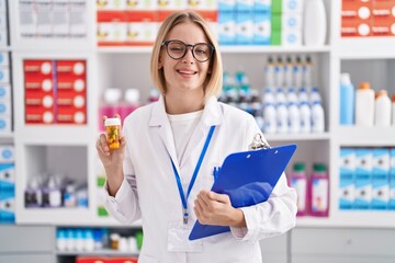 Young caucasian woman working at pharmacy drugstore holding pills winking looking at the camera...