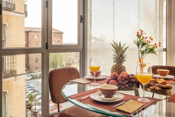 Dining room with a circular glass table full of assorted fruits, glasses of orange juice and brown chairs next to a bay window with a view
