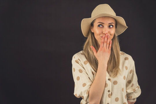 Caucasian blond woman wearing wide sun hat and polka-dot shirt covering mouth with hand looking to the right. Silly suspiscious expression. Dark background studio shot. High quality photo