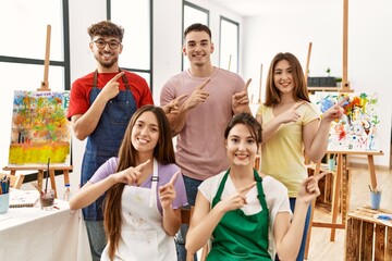 Group of five hispanic artists at art studio smiling and looking at the camera pointing with two hands and fingers to the side.