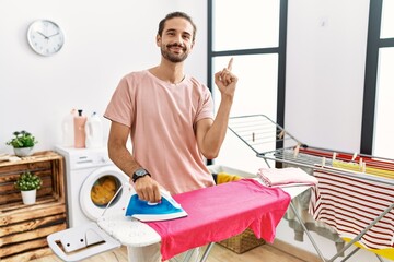 Obraz premium Young hispanic man ironing clothes at home with a big smile on face, pointing with hand finger to the side looking at the camera.