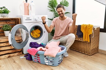 Young hispanic man putting dirty laundry into washing machine very happy and excited doing winner gesture with arms raised, smiling and screaming for success. celebration concept.