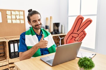 Young hispanic man watching football on laptop cheering game at the office smiling happy and positive, thumb up doing excellent and approval sign