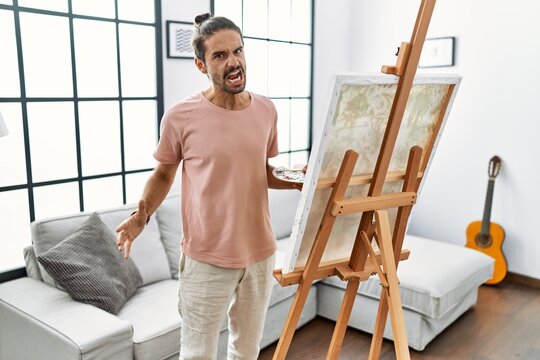 Young hispanic man with beard painting on canvas at home angry and mad screaming frustrated and furious, shouting with anger. rage and aggressive concept.