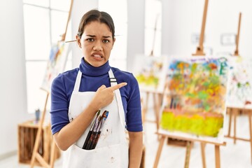 Young brunette woman at art studio pointing aside worried and nervous with forefinger, concerned...