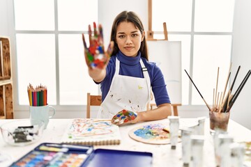 Young brunette woman at art studio with painted hands doing stop sing with palm of the hand....