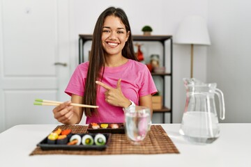 Obraz na płótnie Canvas Young brunette woman eating sushi using chopsticks cheerful with a smile on face pointing with hand and finger up to the side with happy and natural expression
