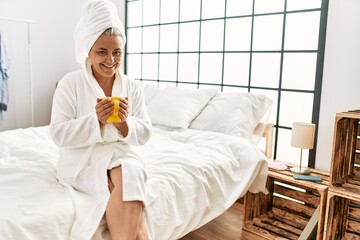 Middle age grey-haired woman drinking cup of coffee sitting on the bed at bedroom.