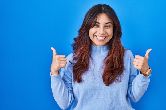 Hispanic young woman standing over blue background success sign doing positive gesture with hand, thumbs up smiling and happy. cheerful expression and winner gesture.