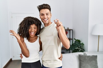 Young interracial couple holding keys of new home celebrating victory with happy smile and winner expression with raised hands