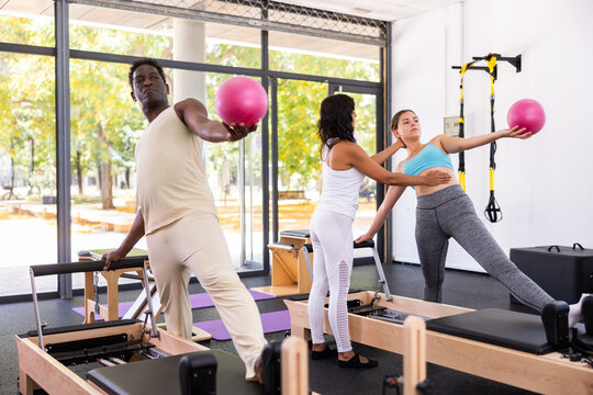 African-american man and caucasian young woman doing exercises at pilates reformers, using mini balls. Hispanic woman trainer correcting their moves.