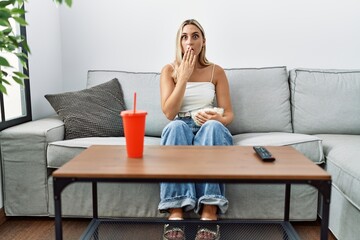 Young blonde woman eating popcorn sitting on the sofa covering mouth with hand, shocked and afraid...