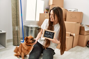 Young hispanic woman holding new home blackboard sitting on floor with dog at new home