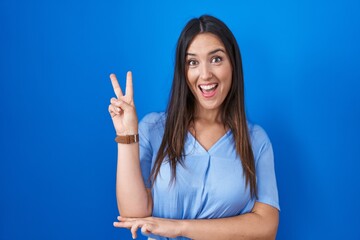 Young brunette woman standing over blue background smiling with happy face winking at the camera...