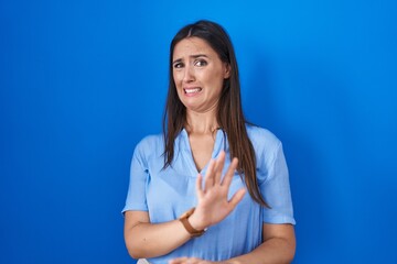 Young brunette woman standing over blue background disgusted expression, displeased and fearful...